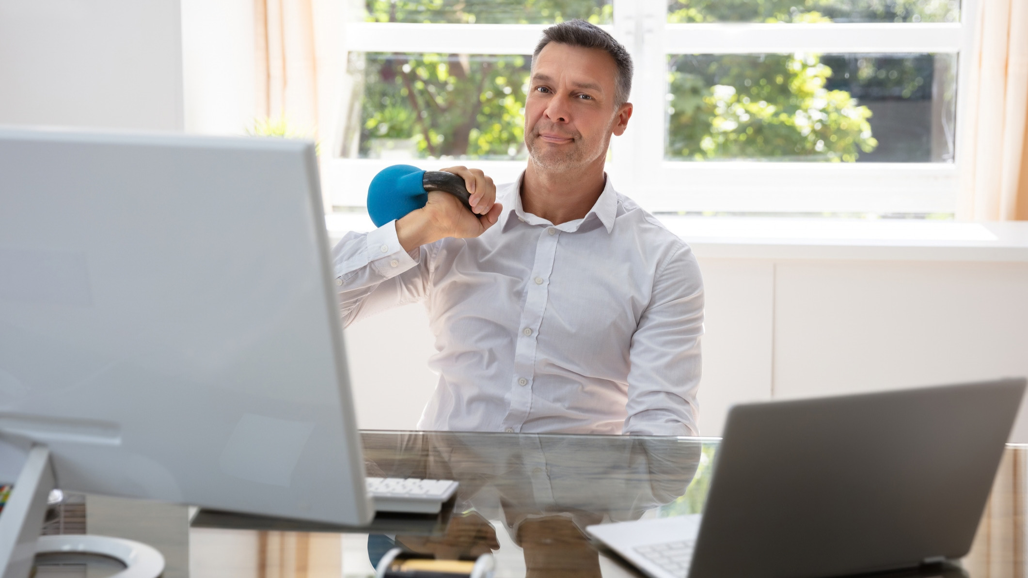 Man sits at a desk, holding a kettlebell on one shoulder. 