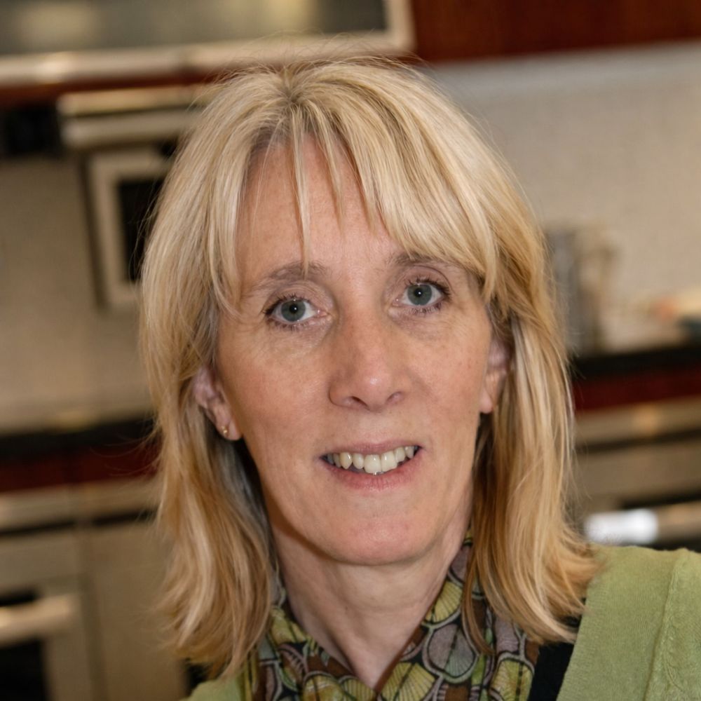 Portrait of a middle aged blonde woman in a kitchen, she's wearing a green jumper and green scarf