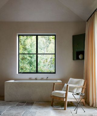 neutral bathroom with limestone tub, white chair and stone floor