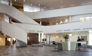 Interior view of the lobby at the Rebobank Westelijke Mijnstreek, Sittard, grey slate tiled floor, wooden stairwells encased in white borders,, glazed front entrance and office space on the 1st and 2nd floors, blurred images of people in the building, woman stood at central desk with chairs, with vase with flowers
