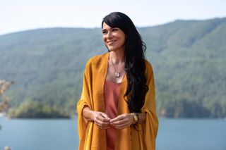 simone kessell as adult lottie wearing an orange shawl standing overlooking a lake in yellowjackets season 2