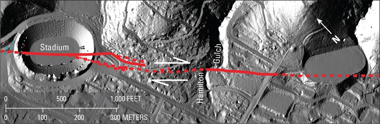 A grayscale aerial image of a football stadium and surrounding streets and low hills. A red line cuts directly through the football stadium from left to right.