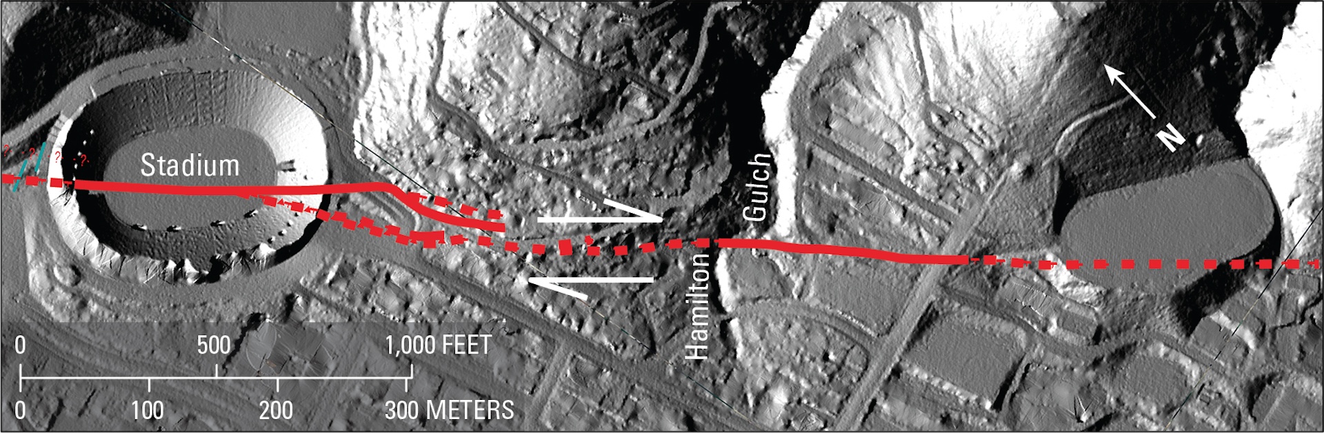 A grayscale aerial image of a football stadium and surrounding streets and low hills. A red line cuts directly through the football stadium from left to right.