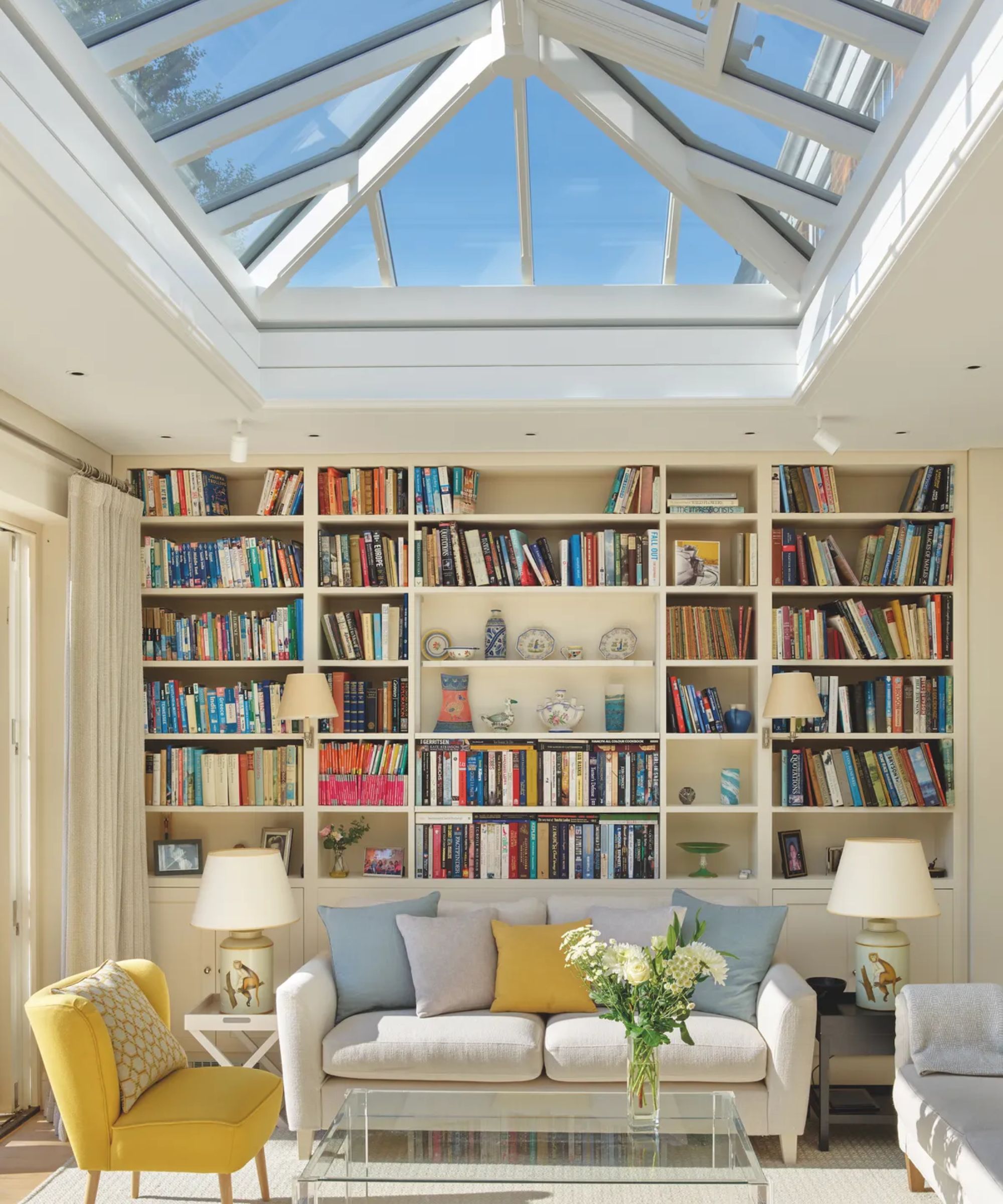 A bright orangery sitting room with a large glass lantern roof flooding light onto a wall of built-in bookshelves, a cream sofa with blue and yellow cushions and a yellow armchair.