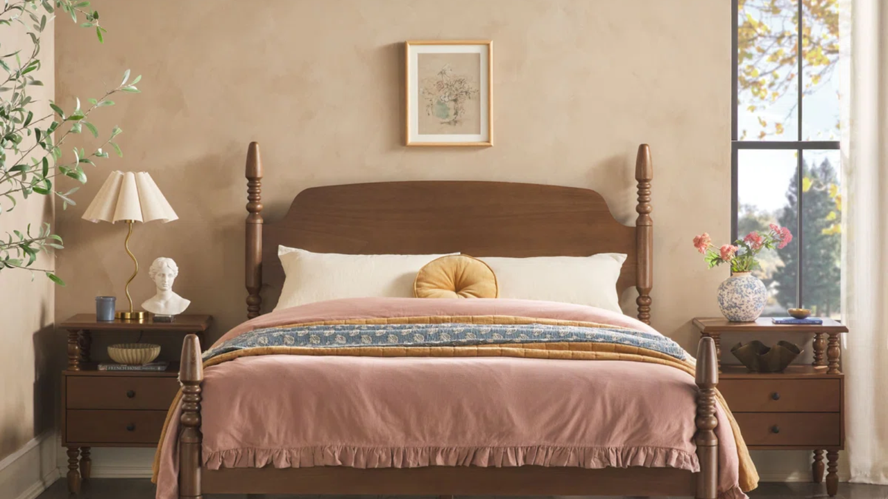 a bedroom with a wooden bed frame, matching wooden side tables, a pink blanket on the bed and a circular yellow pillow in the center, a lamp on the left side table and a vase of florals on the right side table 