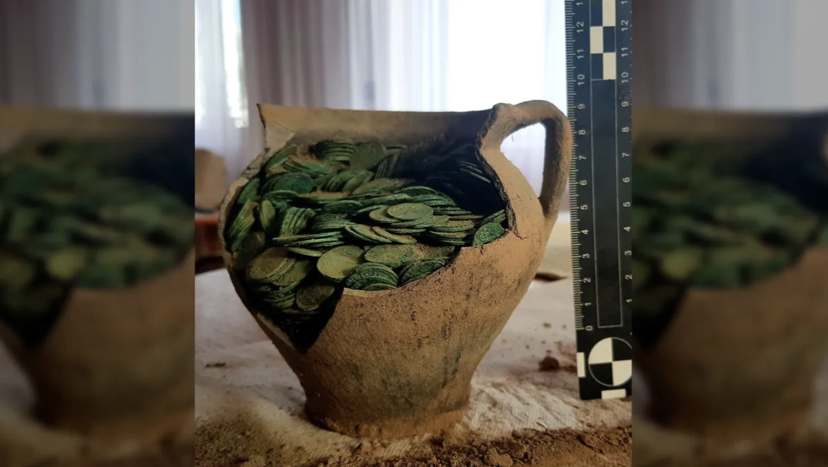 A broken clay jug containing green coins sits on a table.