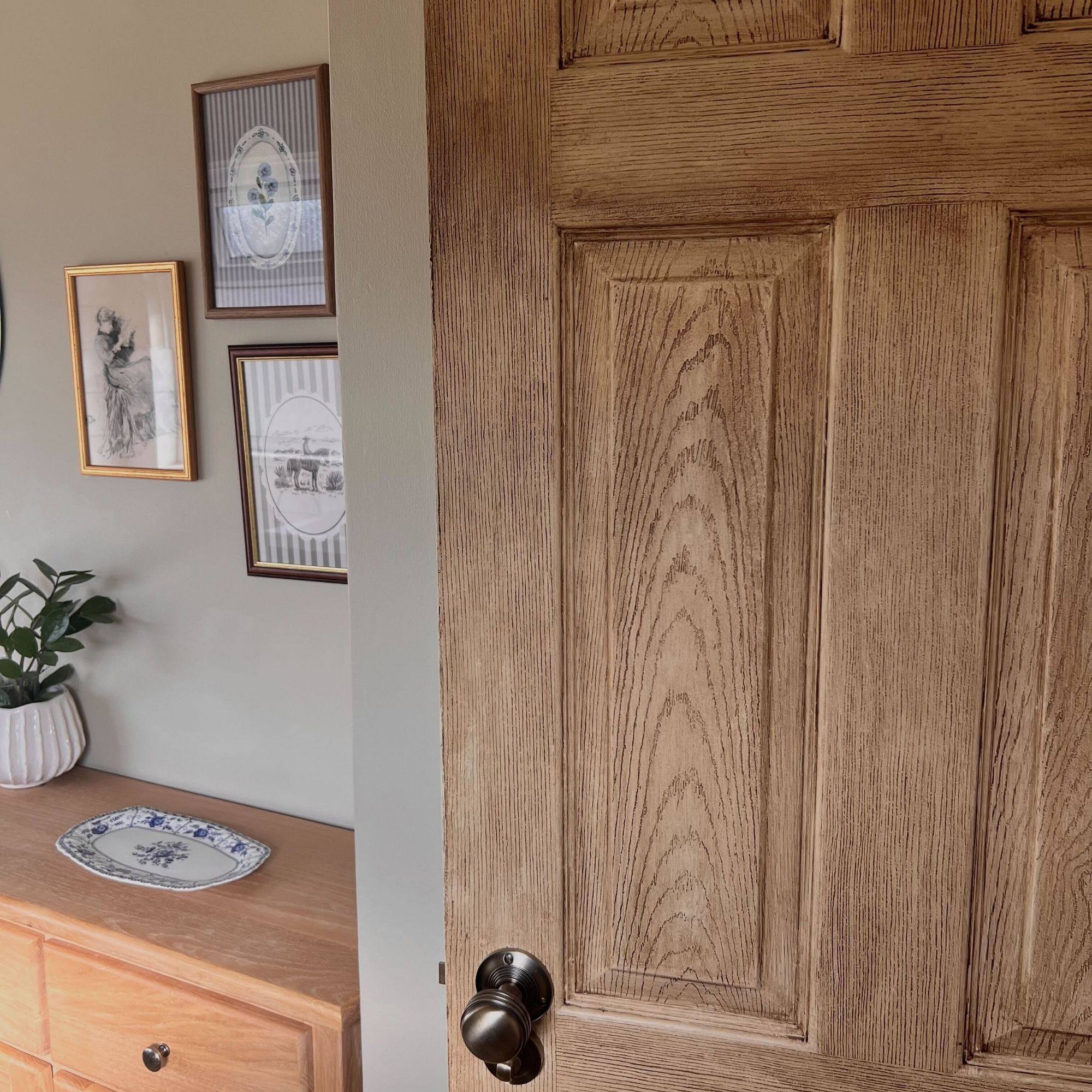 A close-up of an oak door in a brown-grey bathroom with a variety of cottage-style prints on the wall, and a wooden side unit.