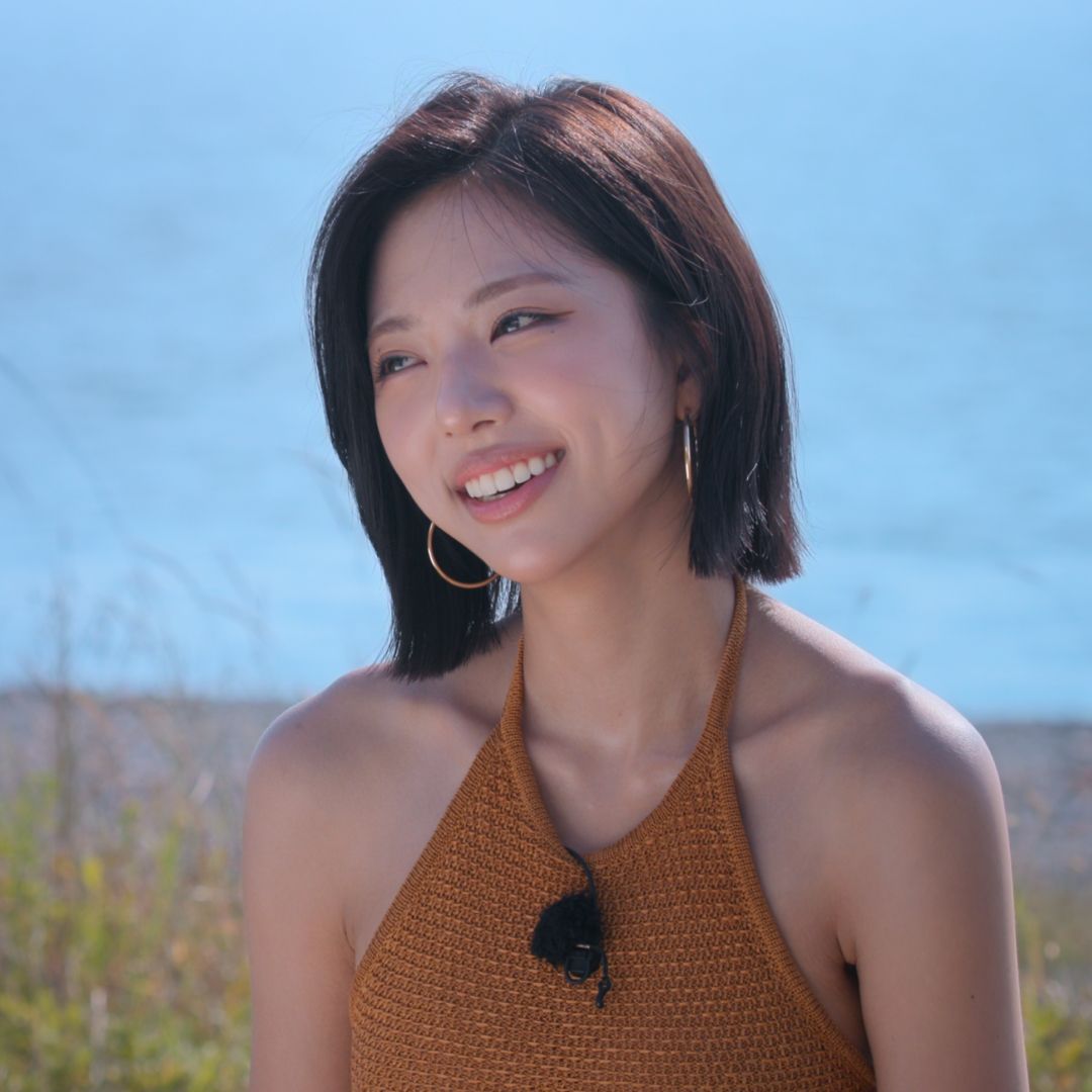A woman wearing a brown halter top and lav mic (model Kim Go-eun) smiles as she sits in front of a small shoreline, in 'Single's Inferno' season 5.