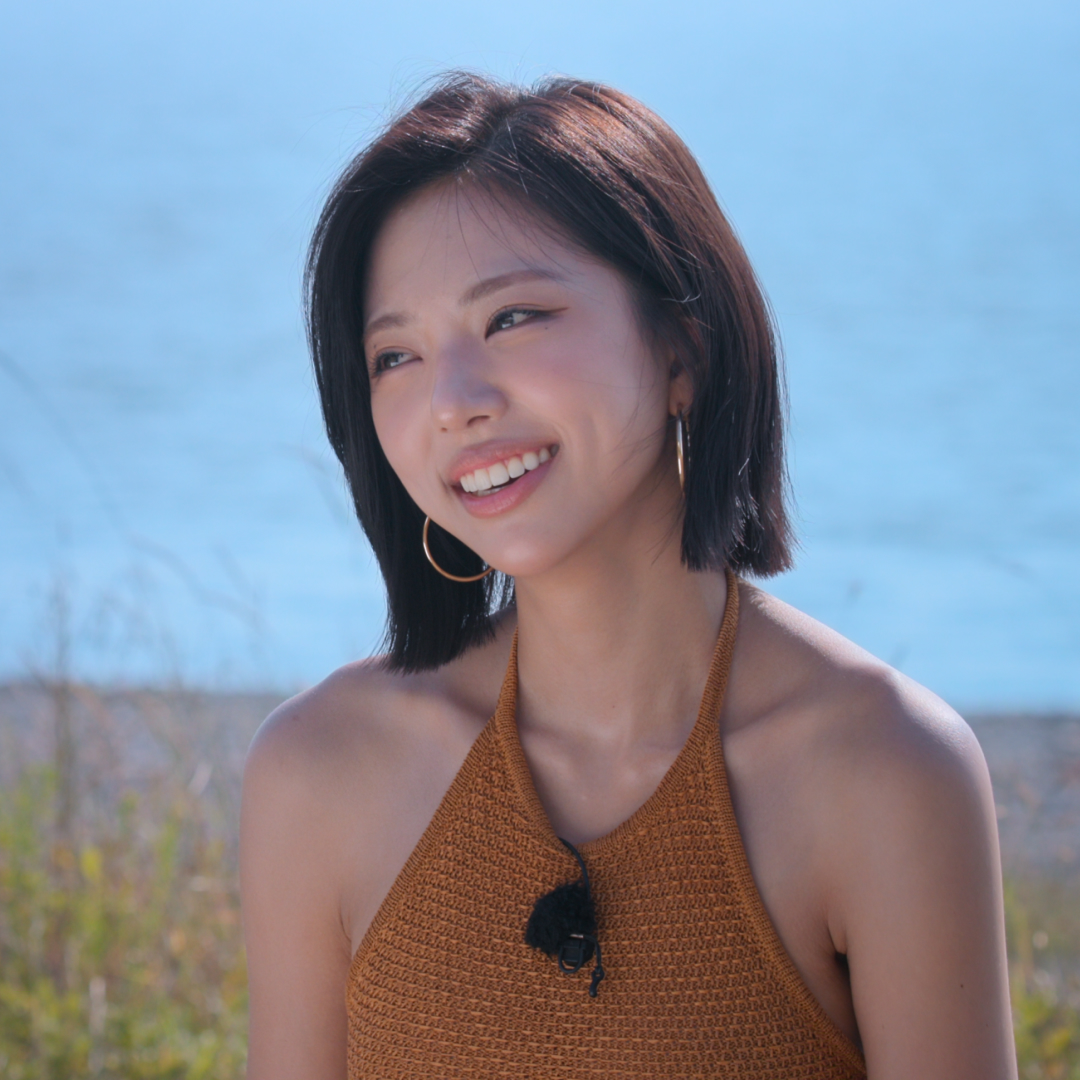 A woman wearing a brown halter top and lav mic (model Kim Go-eun) smiles as she sits in front of a small shoreline, in 'Single's Inferno' season 5.