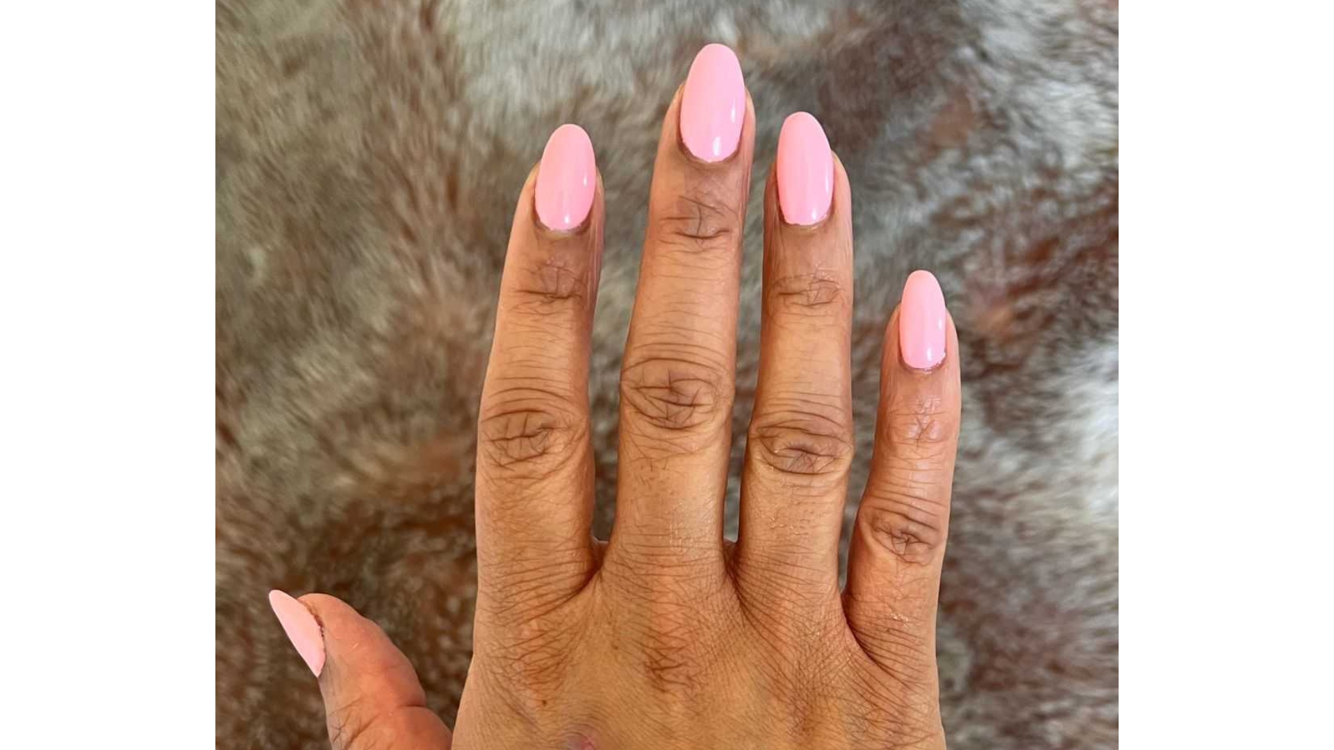 A close-up of Digital Beauty Ecomm Editor, Aleesha Badkar's hand, pictured with long and pink almond nails, in front of a fluffy blanket backdrop