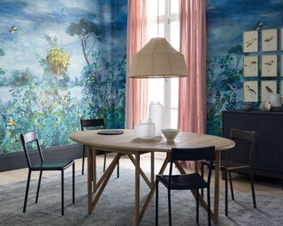 Dining room with striking blue, botanical mural, light wood, oval dining table, four black leather chairs, light wooden flooring with large blue-gray rug, black cabinet with nine bird paintings mounted above, low hanging textured, cream pendant in center of dining table, decorative vases and bowls on table