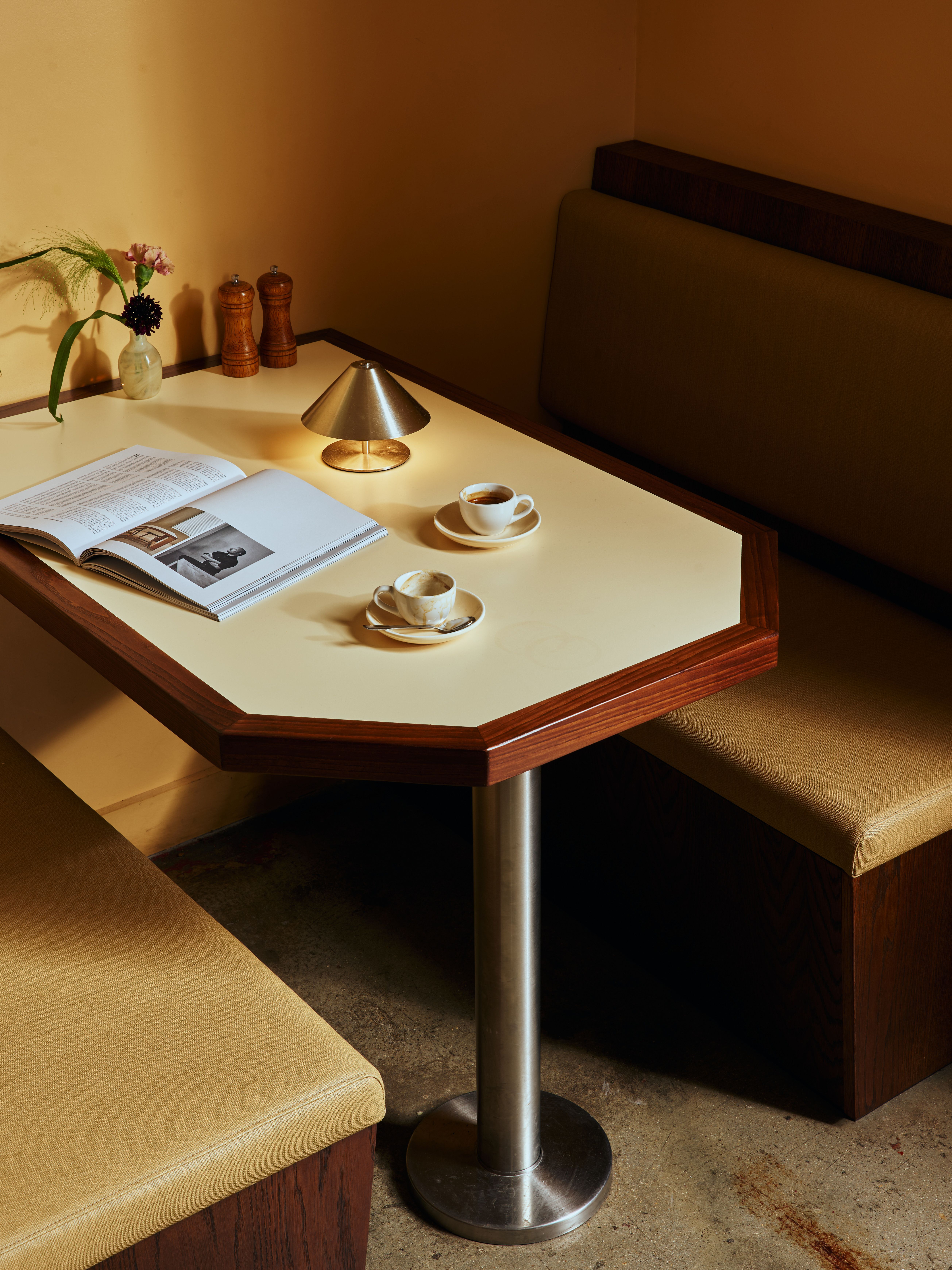 A restaurant banquette booth made of pale, camel-tinted leather, dark wood, and a table crafted with the same materials except for its chrome legs sits in a yellow-painted restaurant with stone floors, topped by two espresso cups, an open book, salt and pepper grinders in wood, a tiny flower vase, and a chrome table lamp.