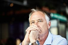 A retired man clasps his hands in front of his mouth and looks into the distance, behind the camera. He appears thoughtful or worried.