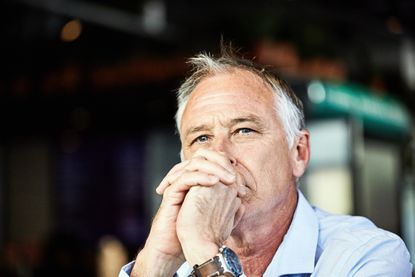 A retired man clasps his hands in front of his mouth and looks into the distance, behind the camera. He appears thoughtful or worried.