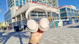 Bose QC Ultra Headphones in white held aloft in an urban outdoors setting