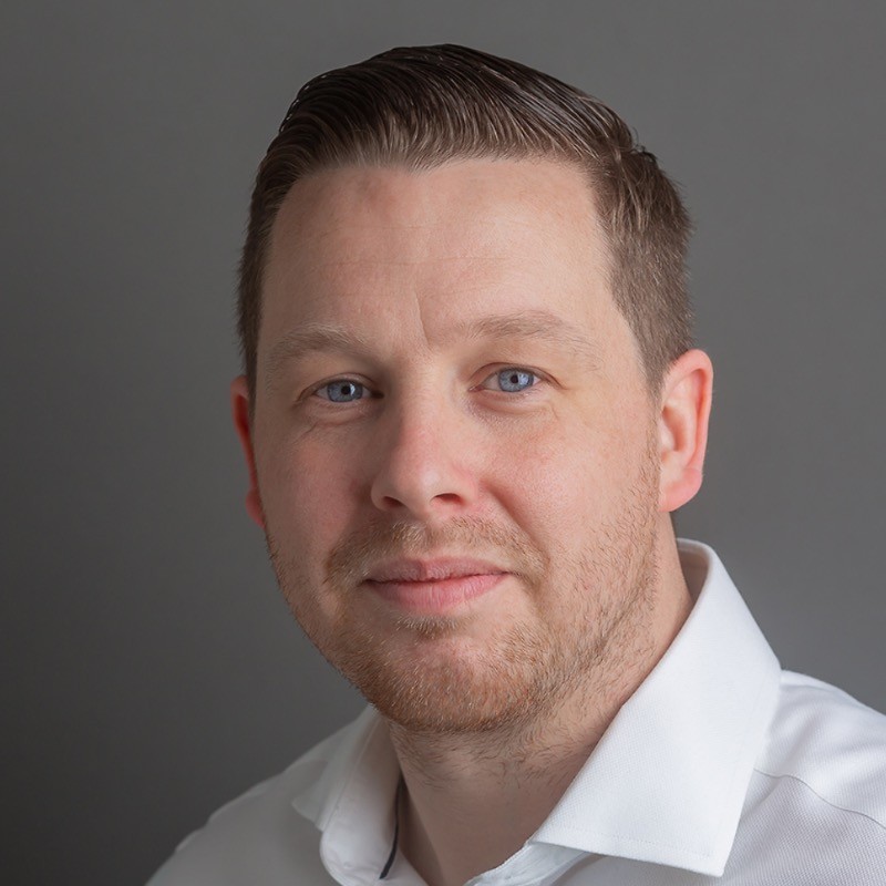 A professional headshot of a man with short dark hair and light stubble wearing a white shirt against a grey background.