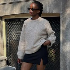 British influencer Liv Madeline poses wearing black retro oval sunglasses, a tan crewneck sweater, and black mini skirt with a thigh slit