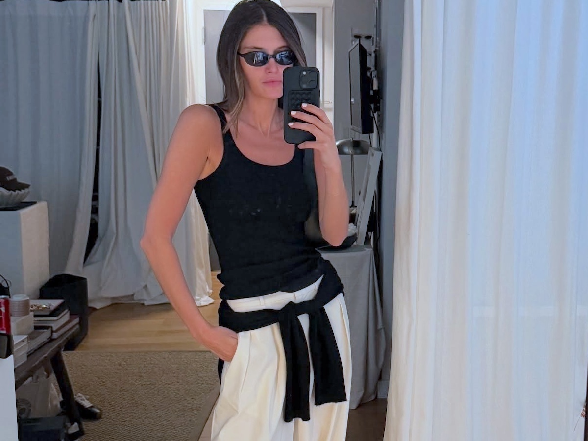 style influencer Coco Schiffer poses for a mirror selfie wearing black oval sunglasses, a black tank top, white trousers, and a black sweater tied around her waist