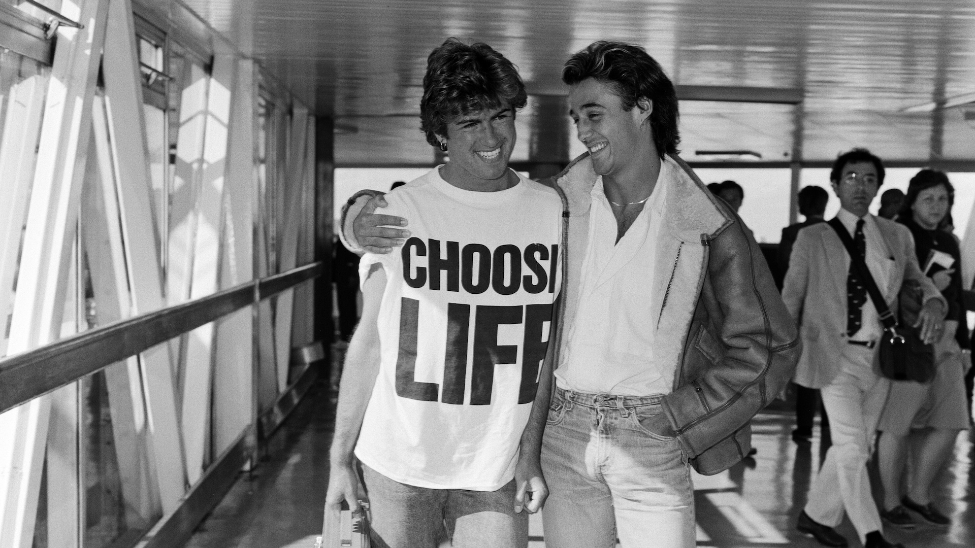 Andrew Ridgeley and George Michael of the pop group Wham!, arriving at London airport. George Michael is wearing a 'Choose Life' t-shirt and holding a portable stereo.