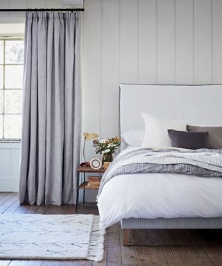 A light gray bedroom curtain with blackout lining in a bedroom with a white bed and white wall panels