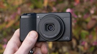 Ricoh GR IV compact camera in user's hand, flower backdrop