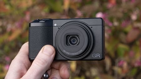 Ricoh GR IV compact camera in user's hand, flower backdrop