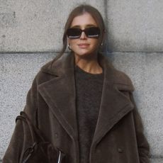 Woman standing against grey wall, wearing brown sweater and brown teddy fall coat, black glasses, and gold hoop earings.