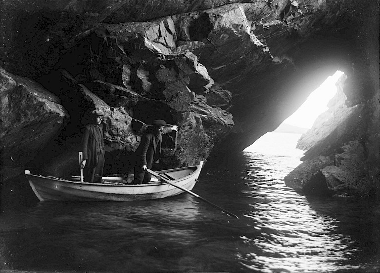 Two men stand in a rowing boat inside a sea cave, with daylight streaming through a rocky opening ahead of them.