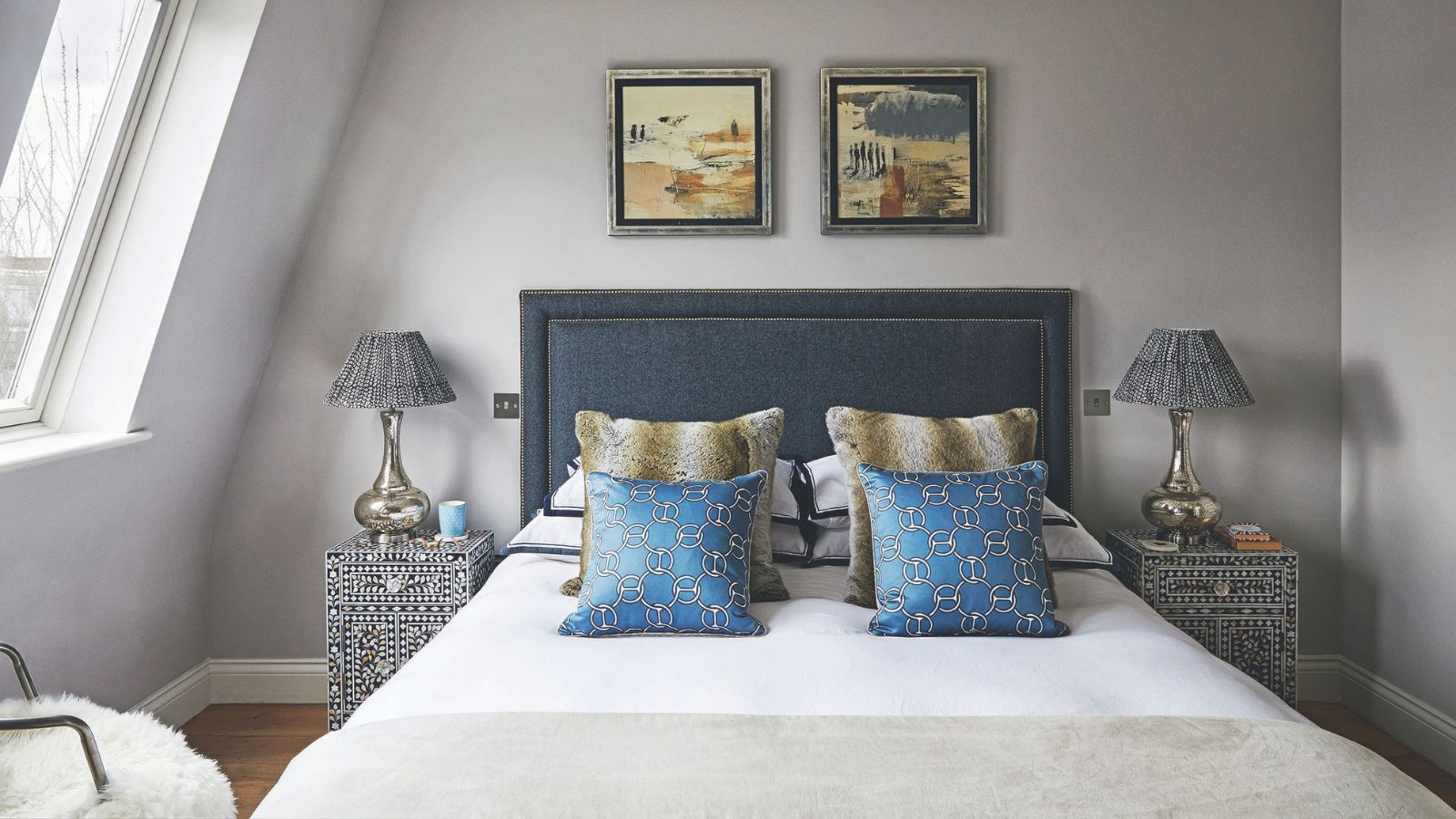 A neutral attic bedroom space with a large double bed with navy upholstered headboard, white bedding, matching cushions, and a matching black and white bedside table on either side with gold lamps with black and white lampshades. Above the headboard are two square landscape artworks.