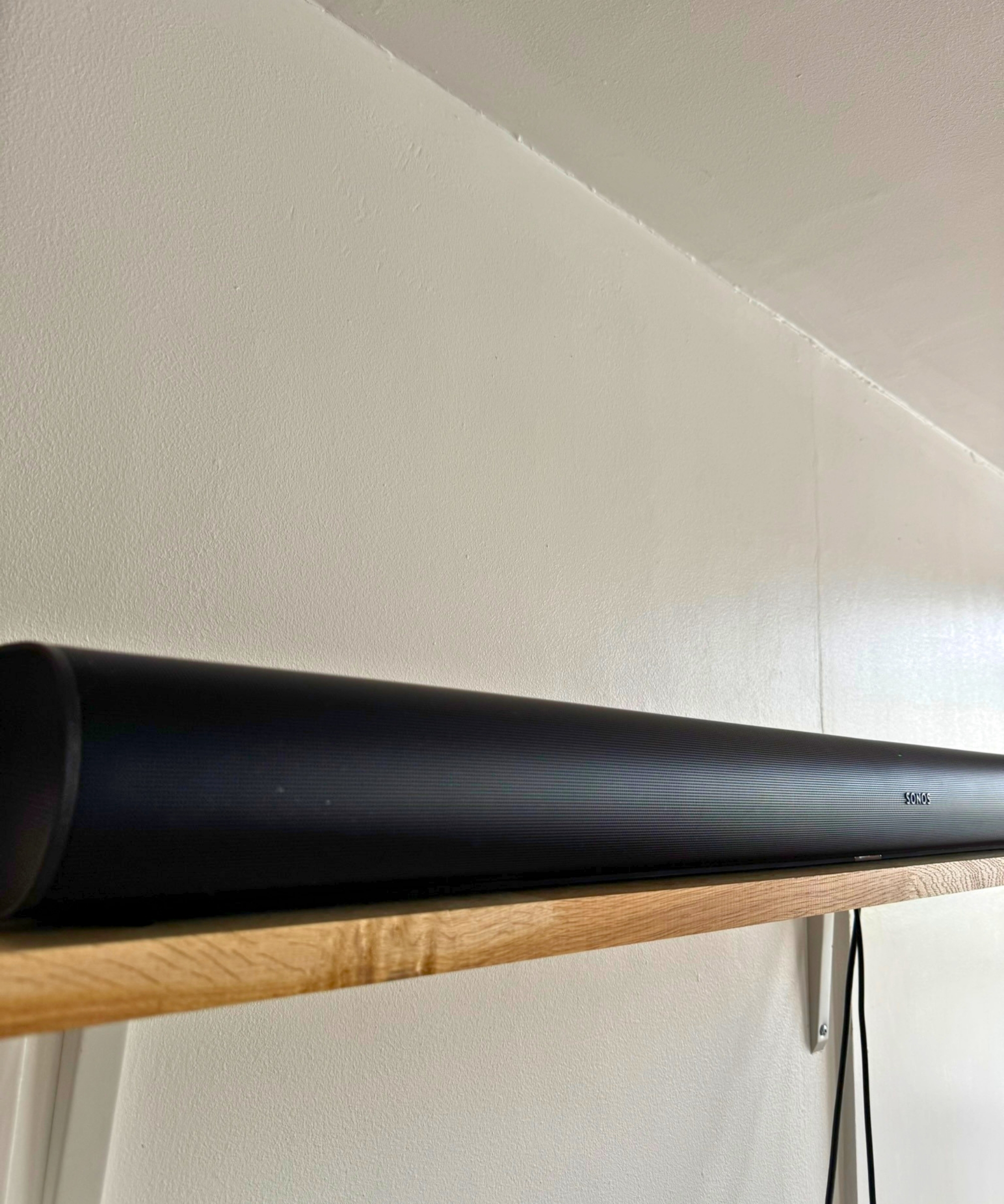 Sonos Arc soundbar on wooden shelf against white wall.