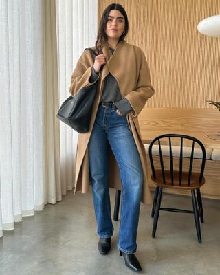 British influencer Anna Newton poses in her chic wood-paneled London kitchen wearing a camel coat, gray sweater, oversize black bag, black belt, jeans, and kitten heel boots.