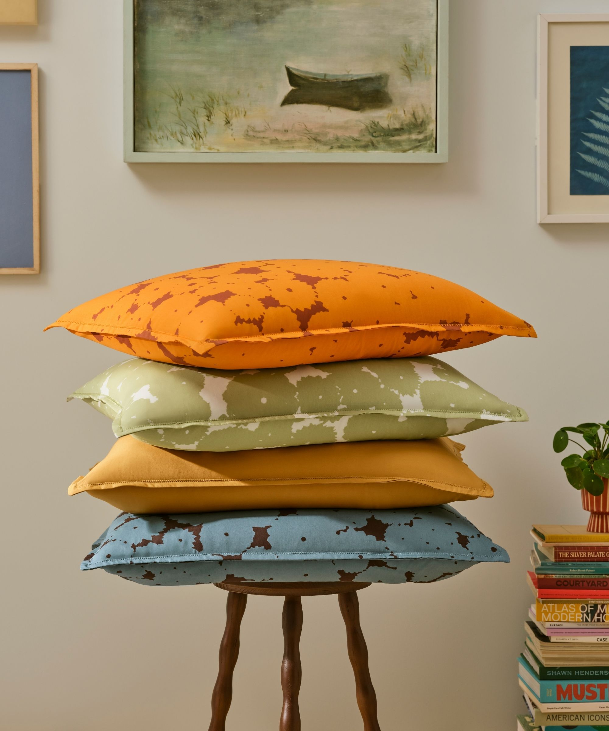 Brooklinen tencel pillowcases on a wooden stool stacked up against a white gallery wall beside a stack of books with a small houseplant on top