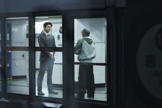 A man in a suit (Gong Yoo) stands in front of a shorter man in a hoodie. They're both seen from the other side of a Seoul subway platform, in 'Squid Game' season 2.