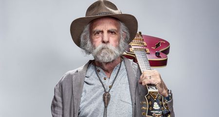 A portait of Bob Weir shot against a light gray background. Weir wears a wide-brimmed hat and has his D'Angelico semi-hollow slung over his shoulder.