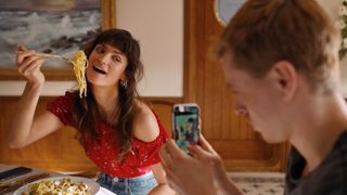 A character from Triangle of Sadness poses for a photo with a mouthful of food, while her boyfriend takes a photo on a phone
