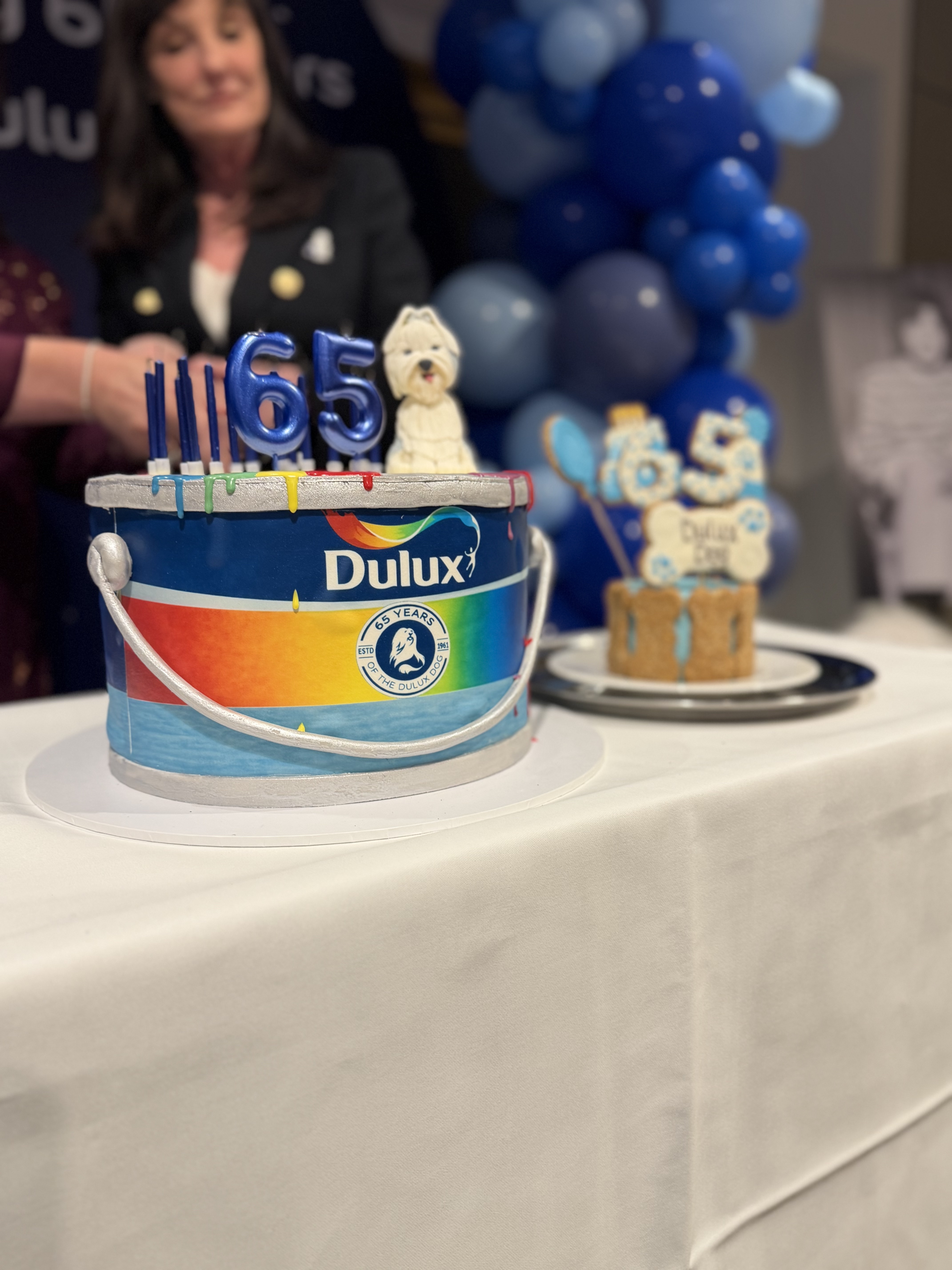 A colourful cake designed to look like a Dulux paint tin sits on a table, topped with blue “65” candles and a small dog figure, with blue balloons and a woman in the background.