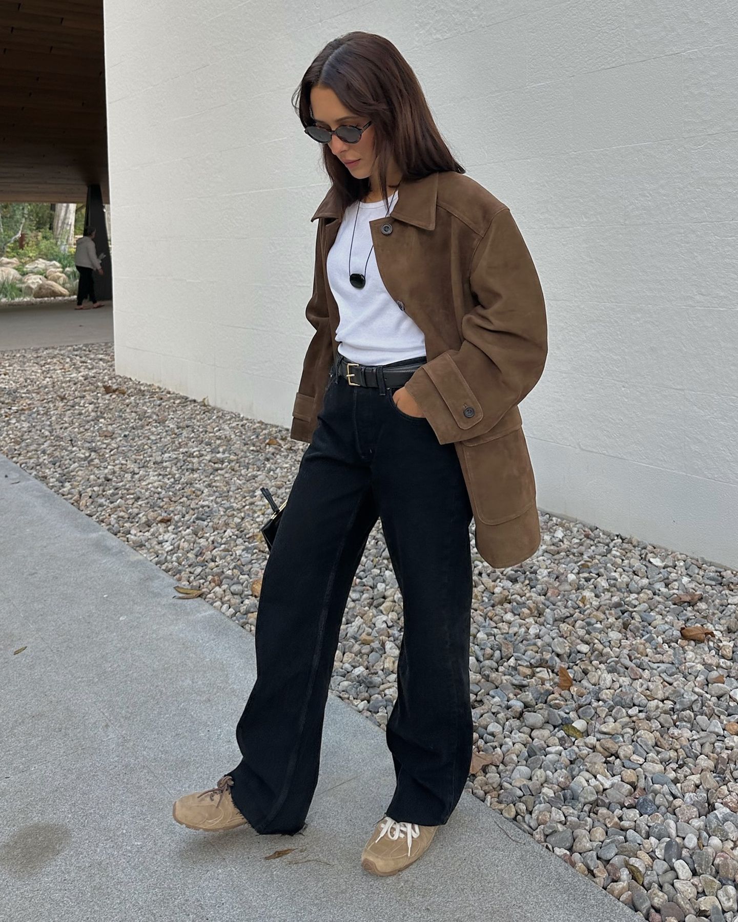 influencer Debora Rosa poses in sunglasses, a pendant cord necklace, brown suede jacket, white tee, black belt, black jeans, and tan brown suede Miu Miu sneakers