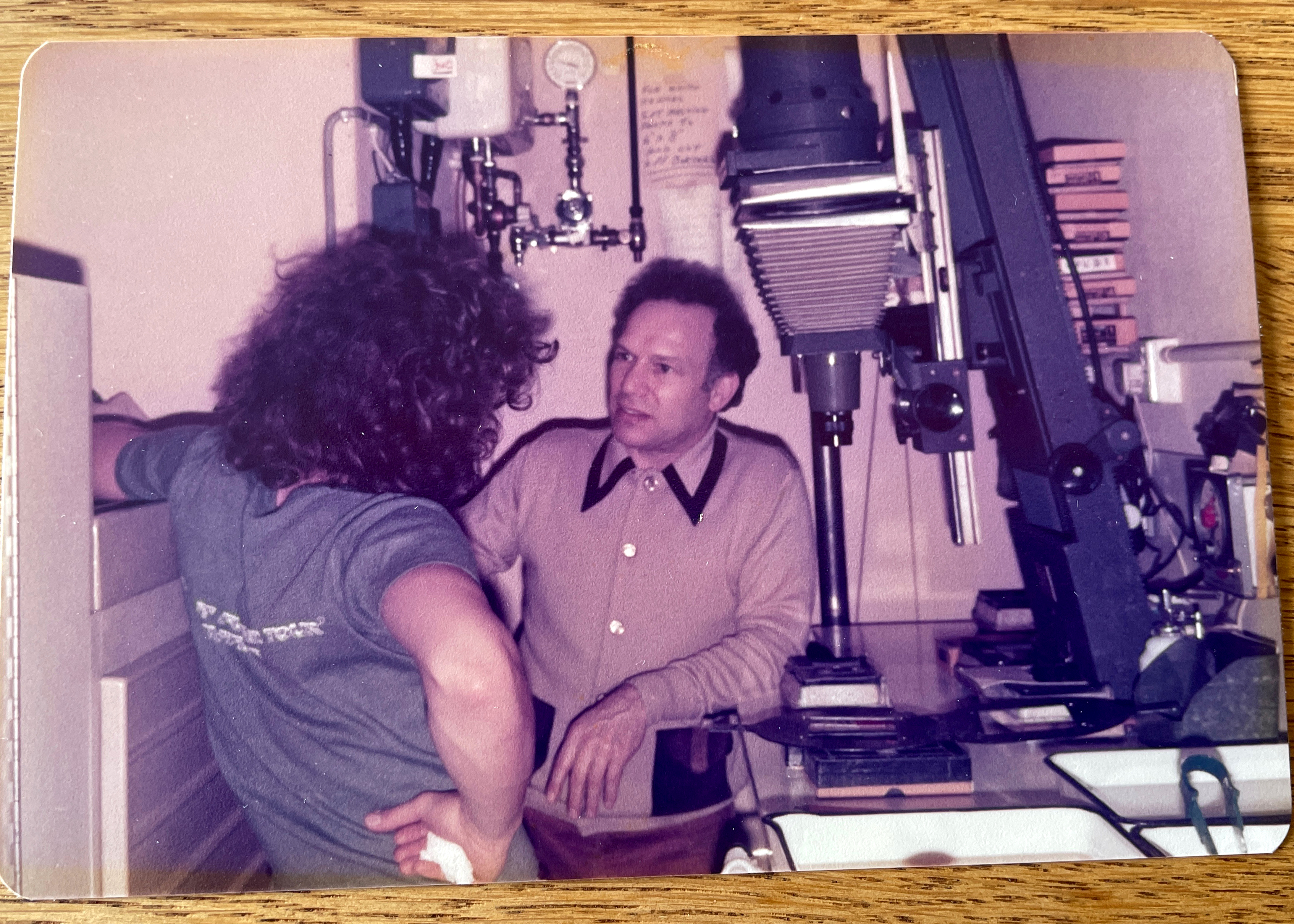 Dave and Philip Sharkey talk in the cramped darkroom at Passport Photo Service in 1978, with a Kodak Versamat processing machine and a Beseler enlarger visible among the equipment around them.
