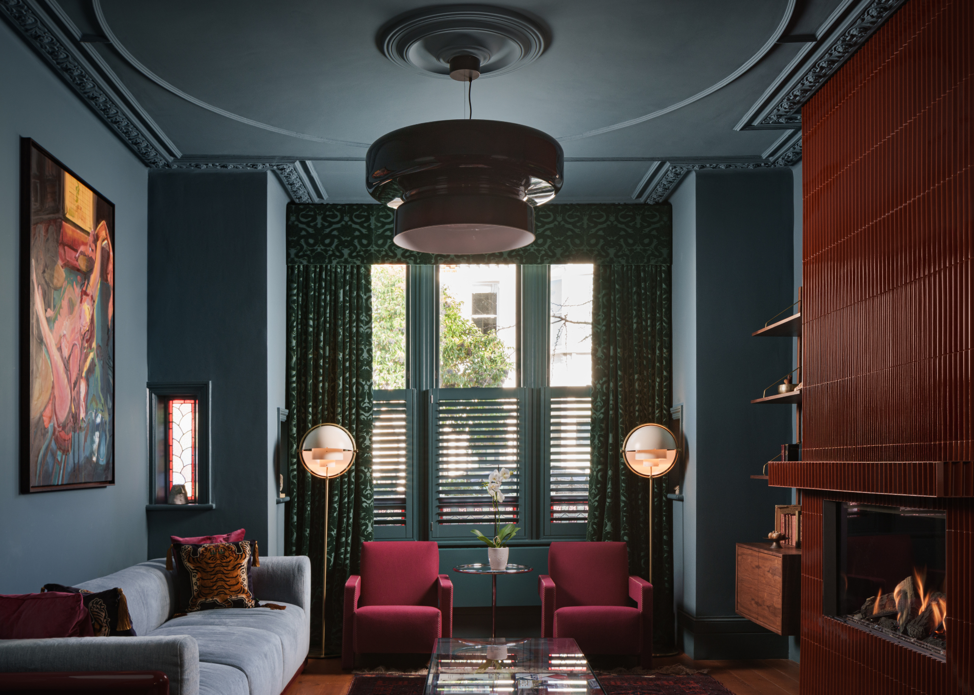 A modern living room with a tiled fireplace, a large artwork on the opposite wall of the fireplace and two accent chairs in the window with round orb floor lamps either side and floor-length green curtains with a pelmet