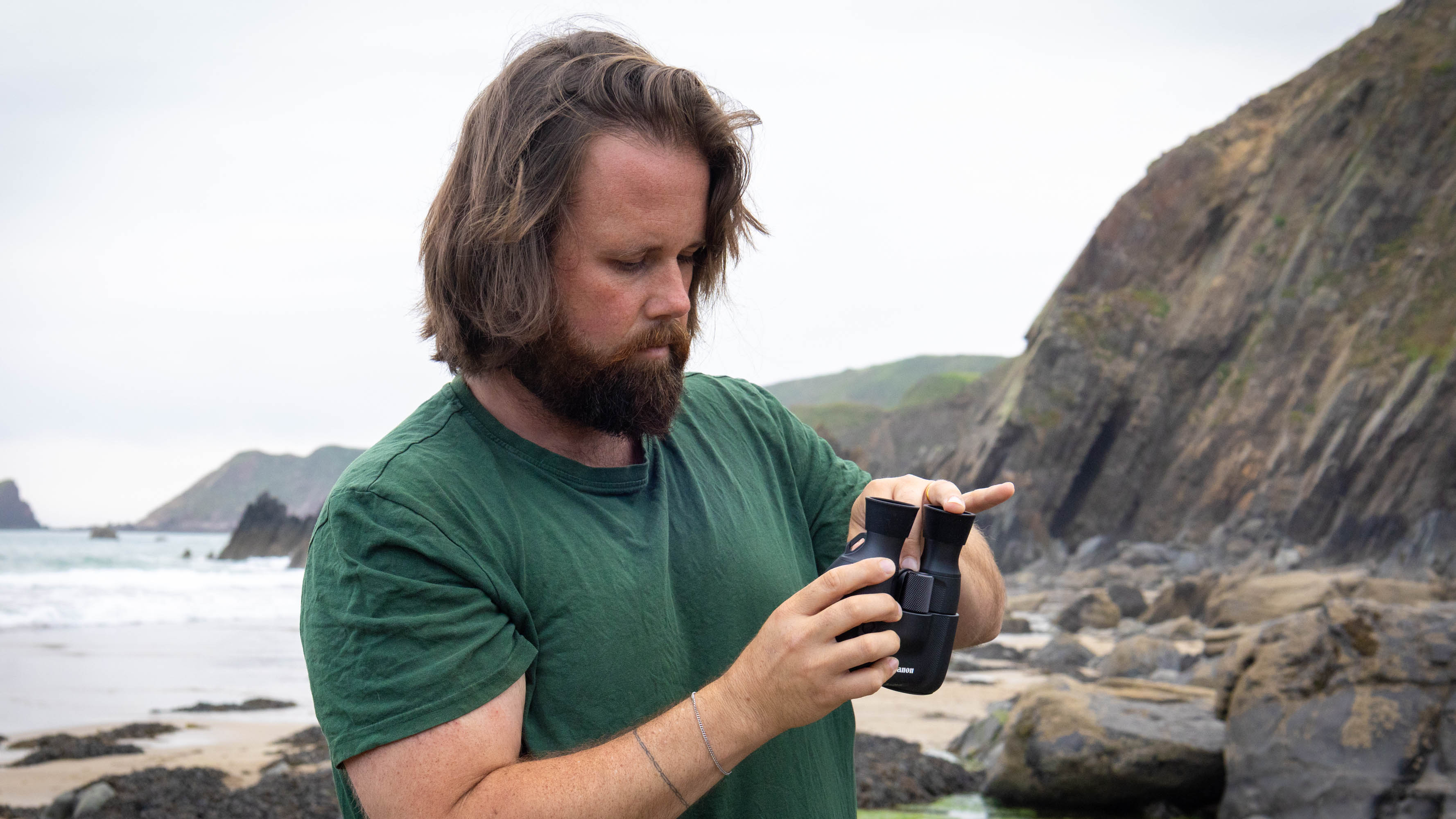 A male holding the Canon 8x20 IS with one hand and accessing another part of the binos with the other. Scene is a beach with coastline.