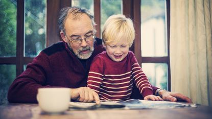 A grandfather and his grandson read a newspaper together at the dining room table.