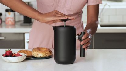 A woman pressing down on a Fellow Clara French press next to a bagel
