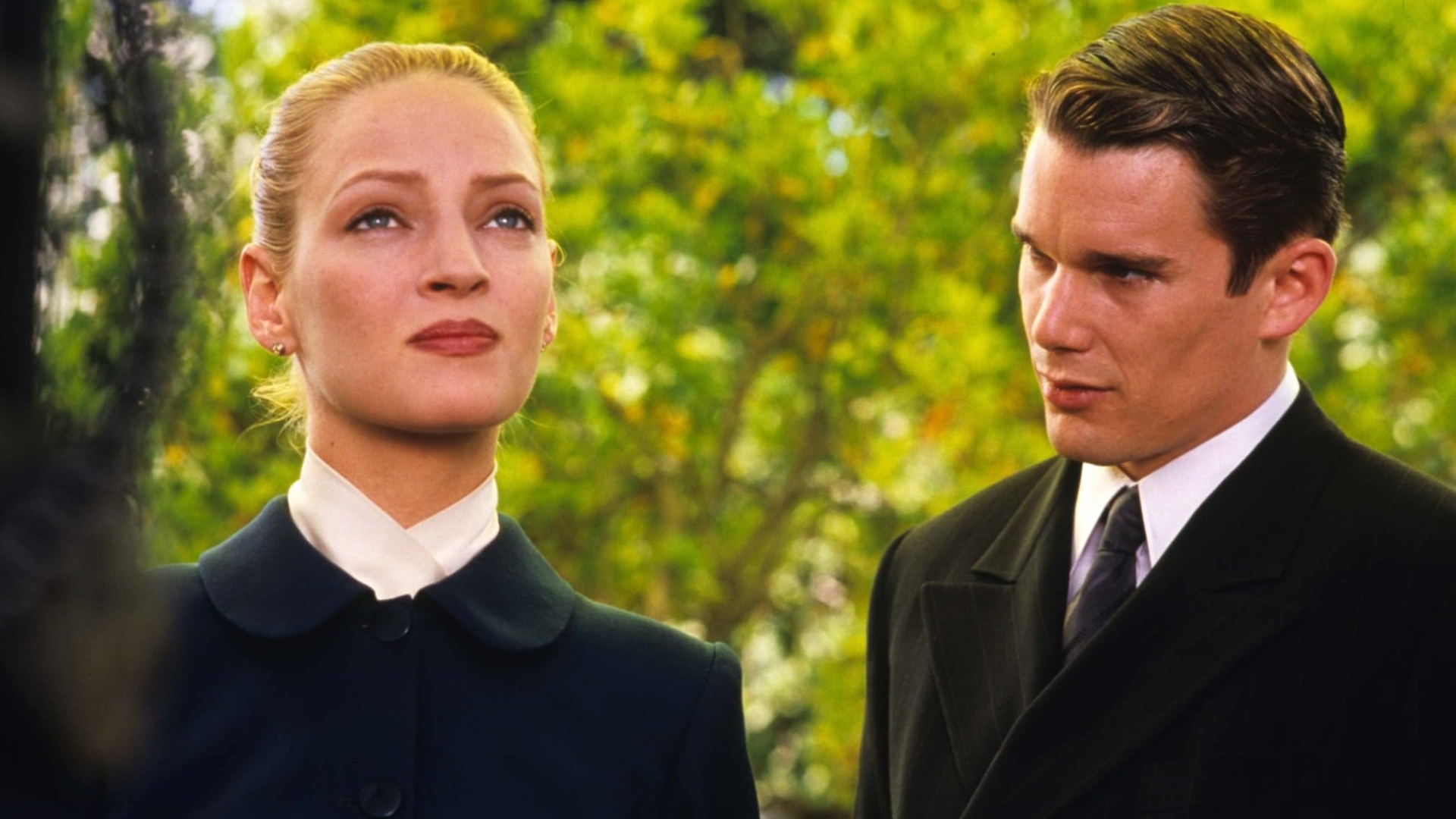 A brown-haired man and blonde woman both dressed in black, stood in a wooded area.