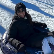 Meghan Markle wearing a black coat and hat sitting in a snow tube and smiling