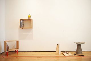 Wooden pieces placed around the room. Wooden box shelf on the white wall. Wooden box shelf with four red legs in the right corner of the room on wooden floor.