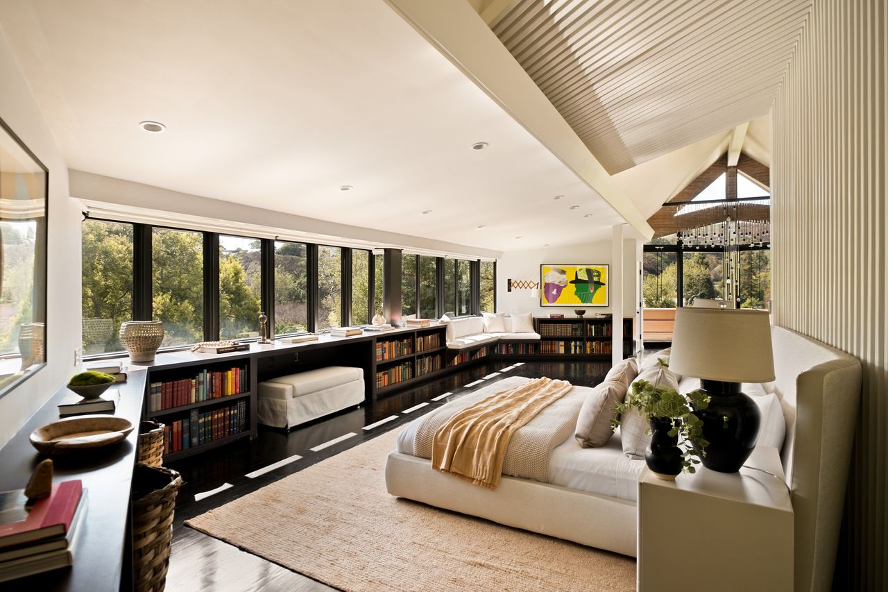 Image of a large bedroom that has long and low bookcases lining the outer wall. About the bookcases are windows that wrap around the room as well. There is a bed in the center and a high white ceiling with wood paneling.