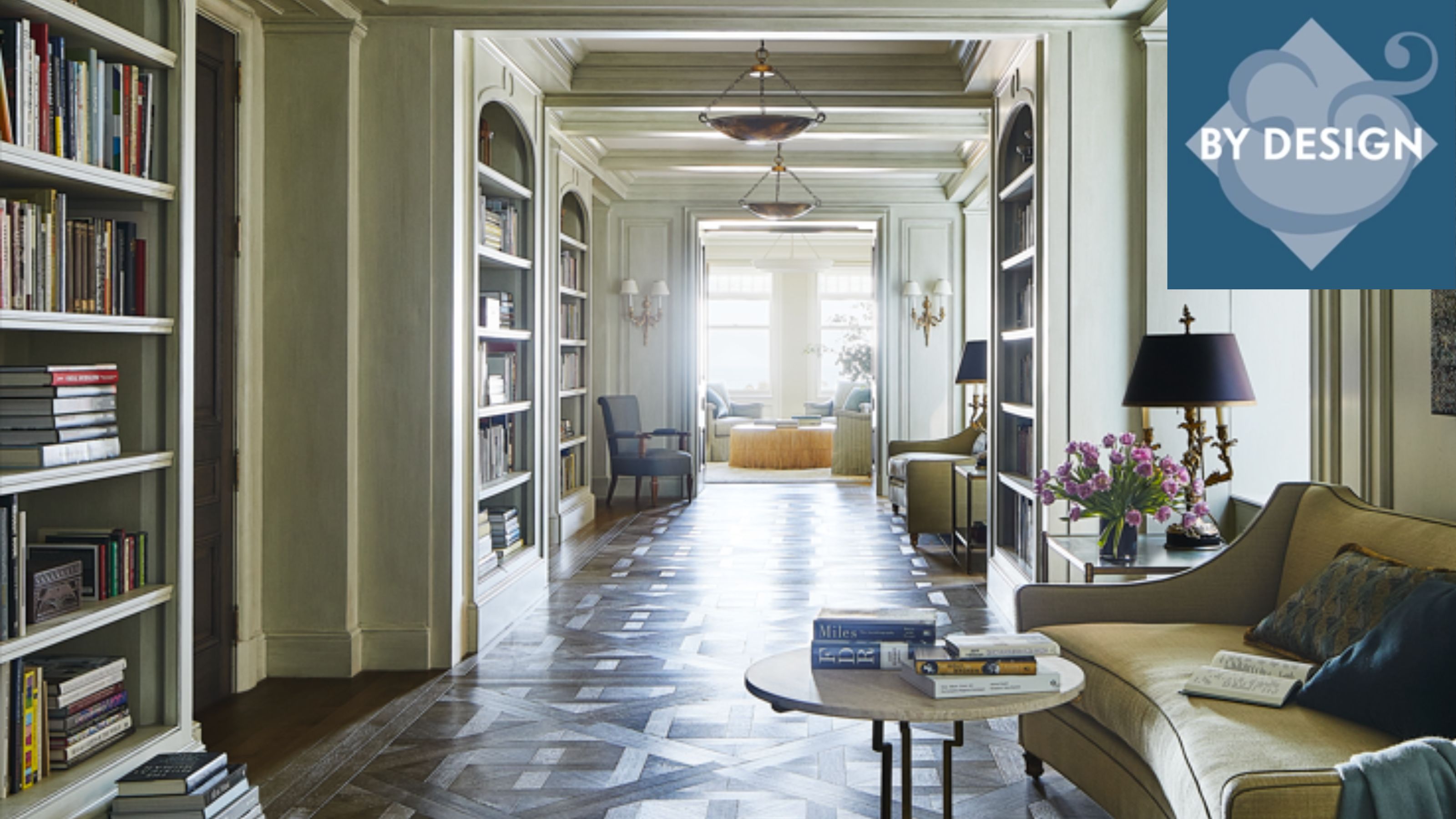 Large, long entryway with bookshelves on either side, wooden flooring, sofa and side table to the lower right hand side of the image, living room with windows in the background