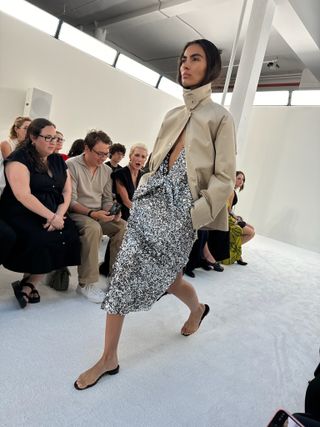 A model at the S/S 25 Brandon Maxwell show wearing a funnel-neck khaki jacket with a silver sequin dress and naked flat sandals.