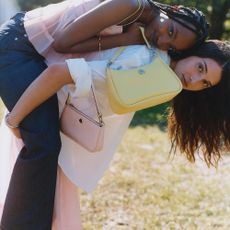 Two models giving eachother a piggyback wearing pastel pink and yellow Kate Spade handbags