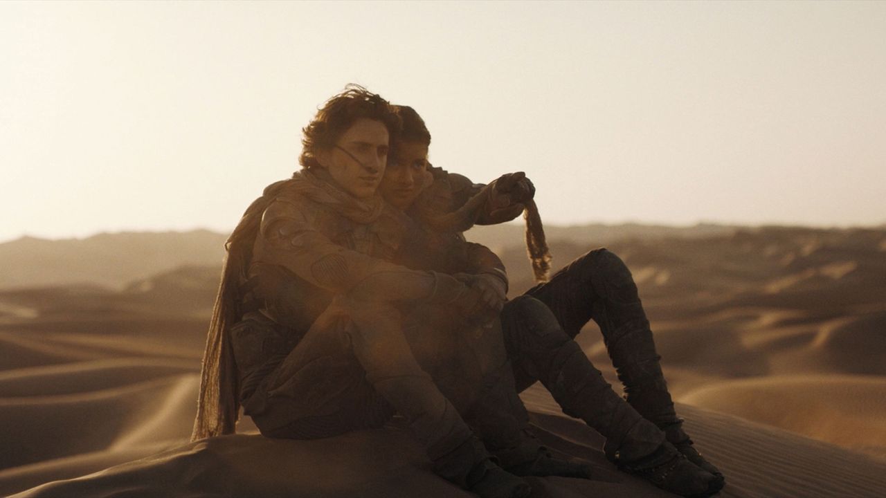 A man and a woman sit on top of a sand dune with a lowering sun in the background.